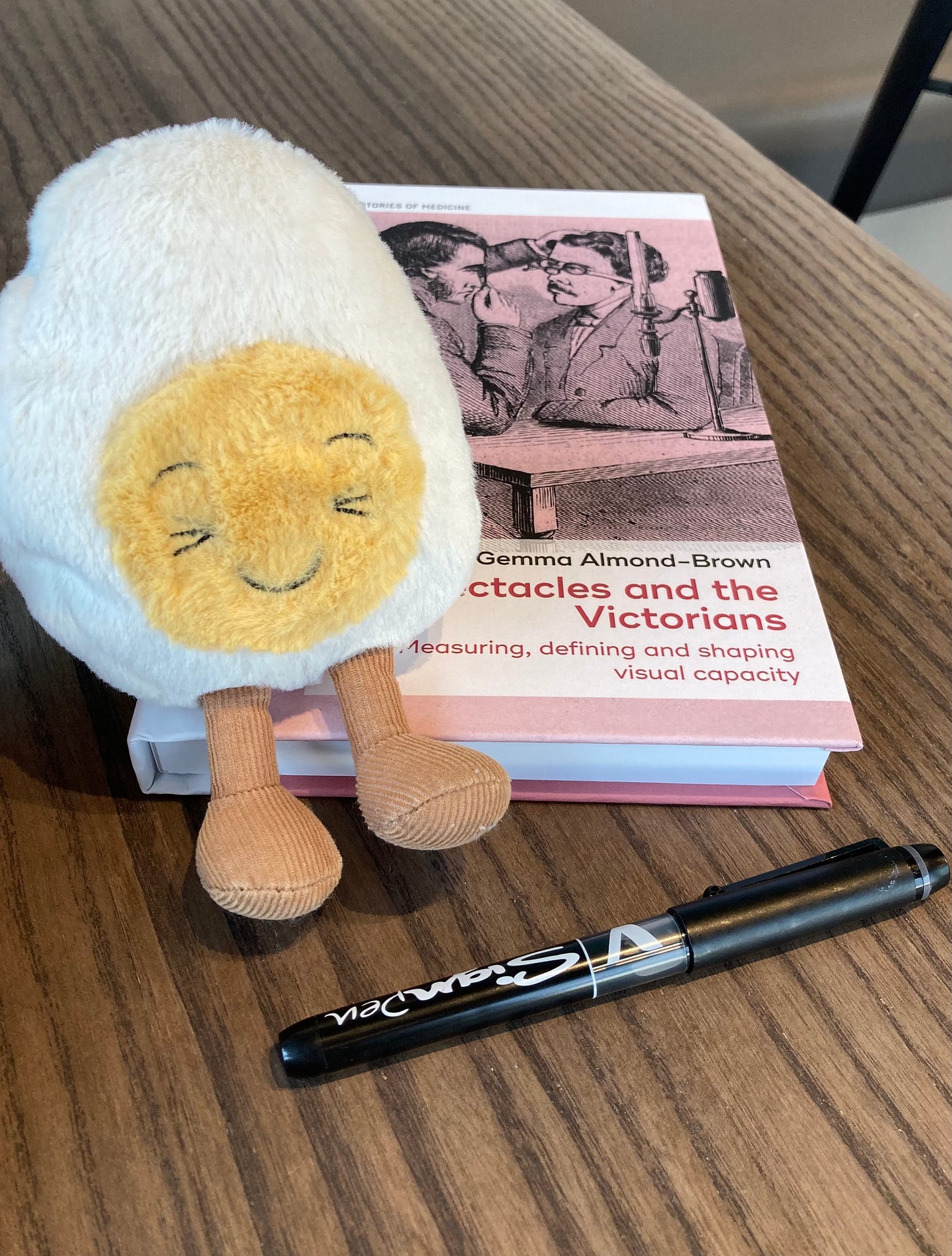 Photograph of Dippy the soft toy egg sitting on top of a hardback book titled Spectacles and the Victorians by Gemma Almond-Brown. They are sitting on a wooden table together, with a inky black pen waiting to be used.