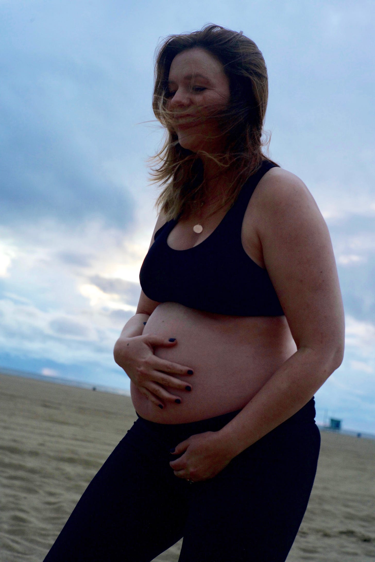 Amber Tamblyn on a beach. The sky is overcast. The wind has blown her hair into her face but you can make out see is laughing. She wears a sports bra and leggings, showing her pregnant stomach which she rests a hand on.