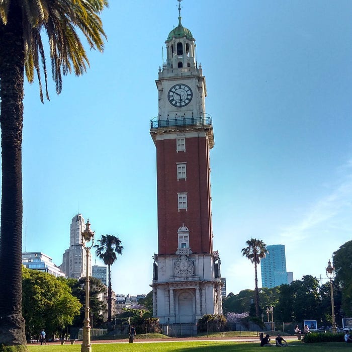 paisagem urbana: praça com várias árvores, a maioria palmeiras. no centro, uma torre de tijolinhos aparentes com 4 janelas, um mirante, um relógio e uma cúpula. ao fundo, vário prédios, com dois em destaque. à esquerda um edifício branco com uma torre sobre ele. à direita, um edifício espelhado.