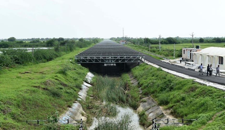 black metal trusses holding solar panels are situated over a grassy canal. Several people walk on a path along the canal.