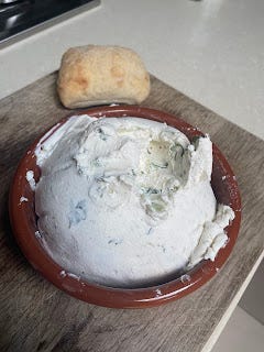 A brown ceramic bowl, sitting on a chopping board, with white, creamy labneh piled high within it. It has flecks of green where herbs are visible.