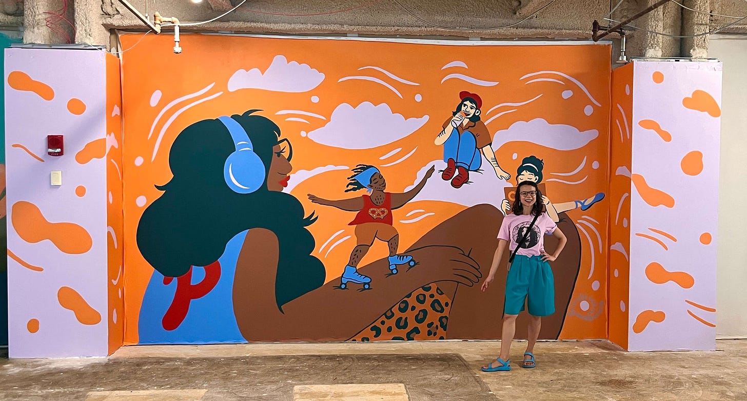 An artist cheesing in front of her completed mural featuring a giant girl listening to music with smaller characters rollerskating, reading and dancing, and eating water ice.