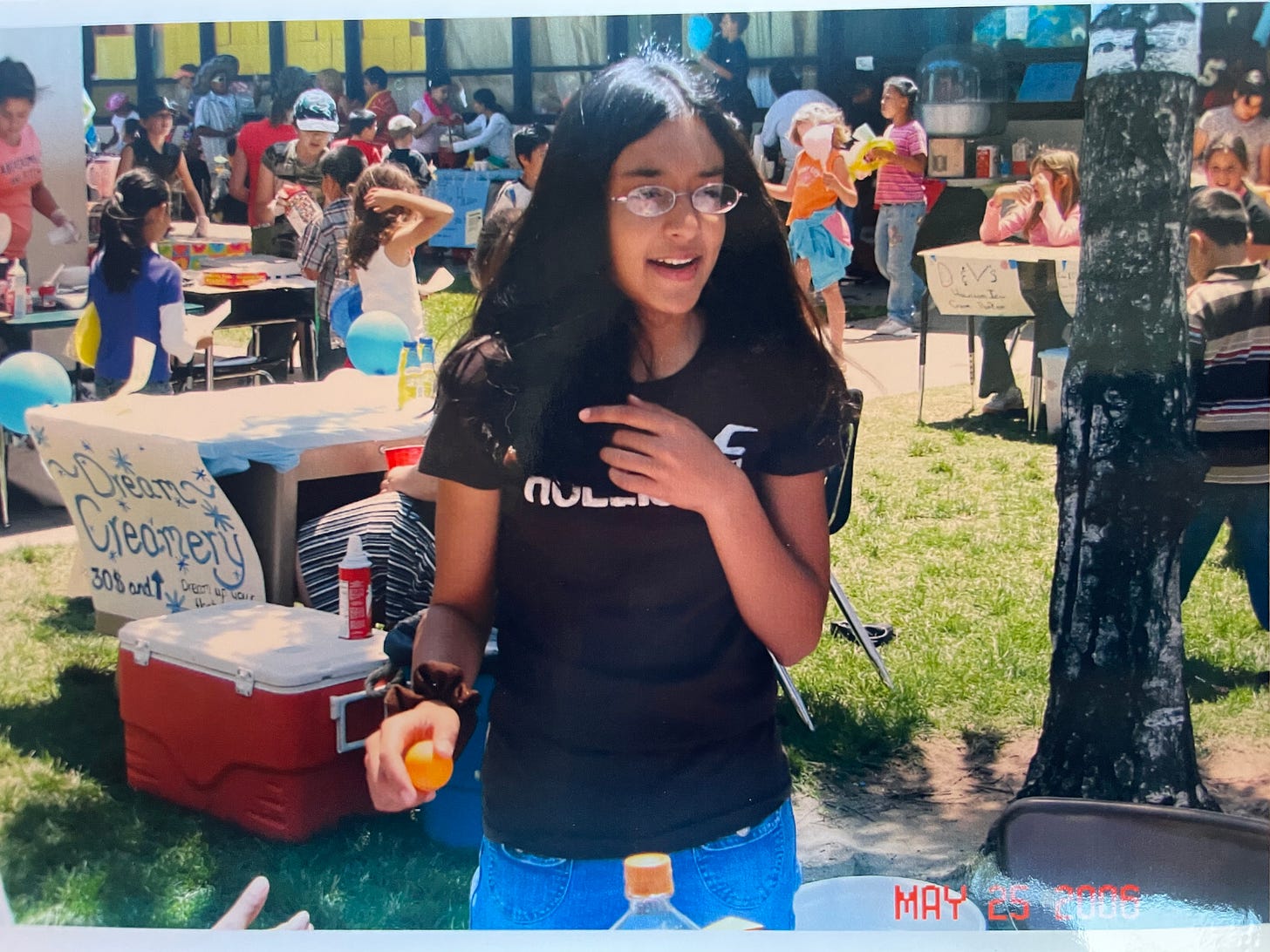 An Indian preteen girl wears a brown Hollister shirt and jeans. She wears glasses and flips her hair, holding an orange in one hand. She appears to be in a school fair setting.
