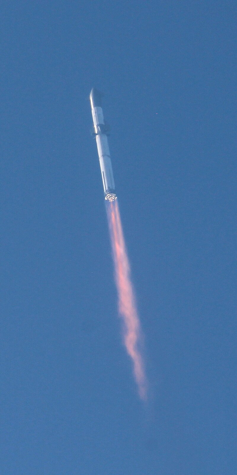 A photo of Starship launching against a blue sky. Flames trail from the rocket, but a handful of the 33 engines do not appear to be lit.