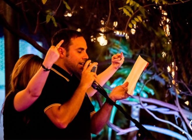 Derrick stands at a microphone, reading from one of his poetry books. Amber stands immediately behind him. Her hands are up near Derrick's head on either side of him as she gives the middle finger. 