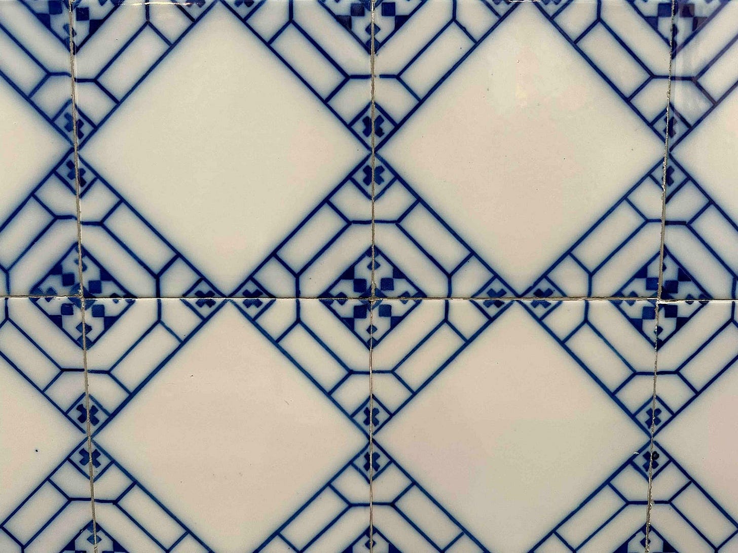 A close-up image of a tiled surface with a repeating pattern. The tiles are square-shaped with white centers and edges decorated in blue geometric and floral designs. The pattern creates a visual effect where blue diamond shapes appear at the intersections of the white squares. There are visible grout lines between the tiles, and slight imperfections can be seen on the surface, indicating wear or age. A close-up image of a tiled surface with a repeating pattern. The tiles are square-shaped with white centers and edges decorated in blue geometric and floral designs. The pattern creates a visual effect where blue diamond shapes appear at the intersections of the white squares. There are visible grout lines between the tiles, and slight imperfections can be seen on the surface, indicating wear or age.