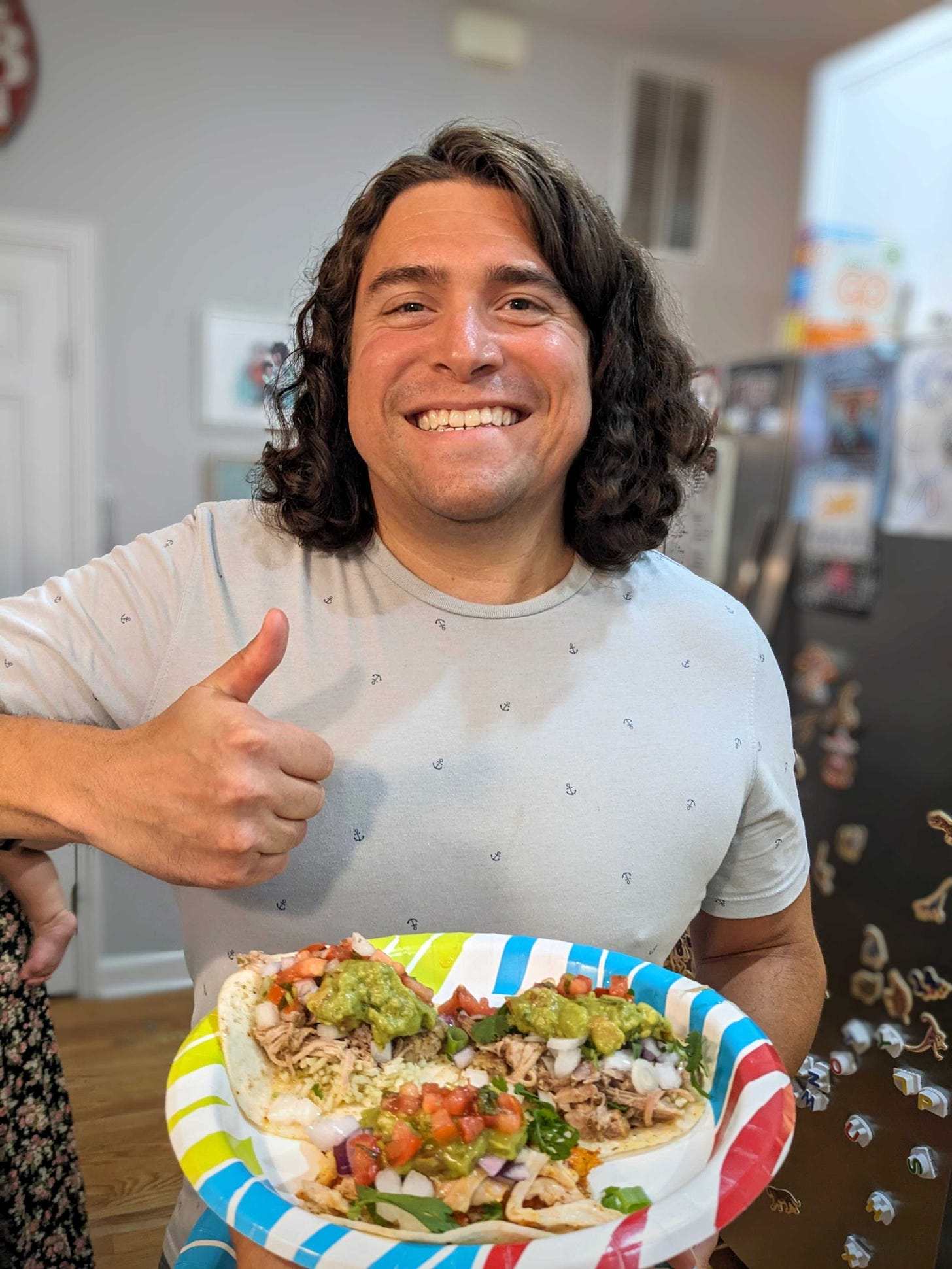 a white man with shoulder-length brown hair and a blue shirt holds a plate of tacos, grinning ear-to-ear and giving thumbs up