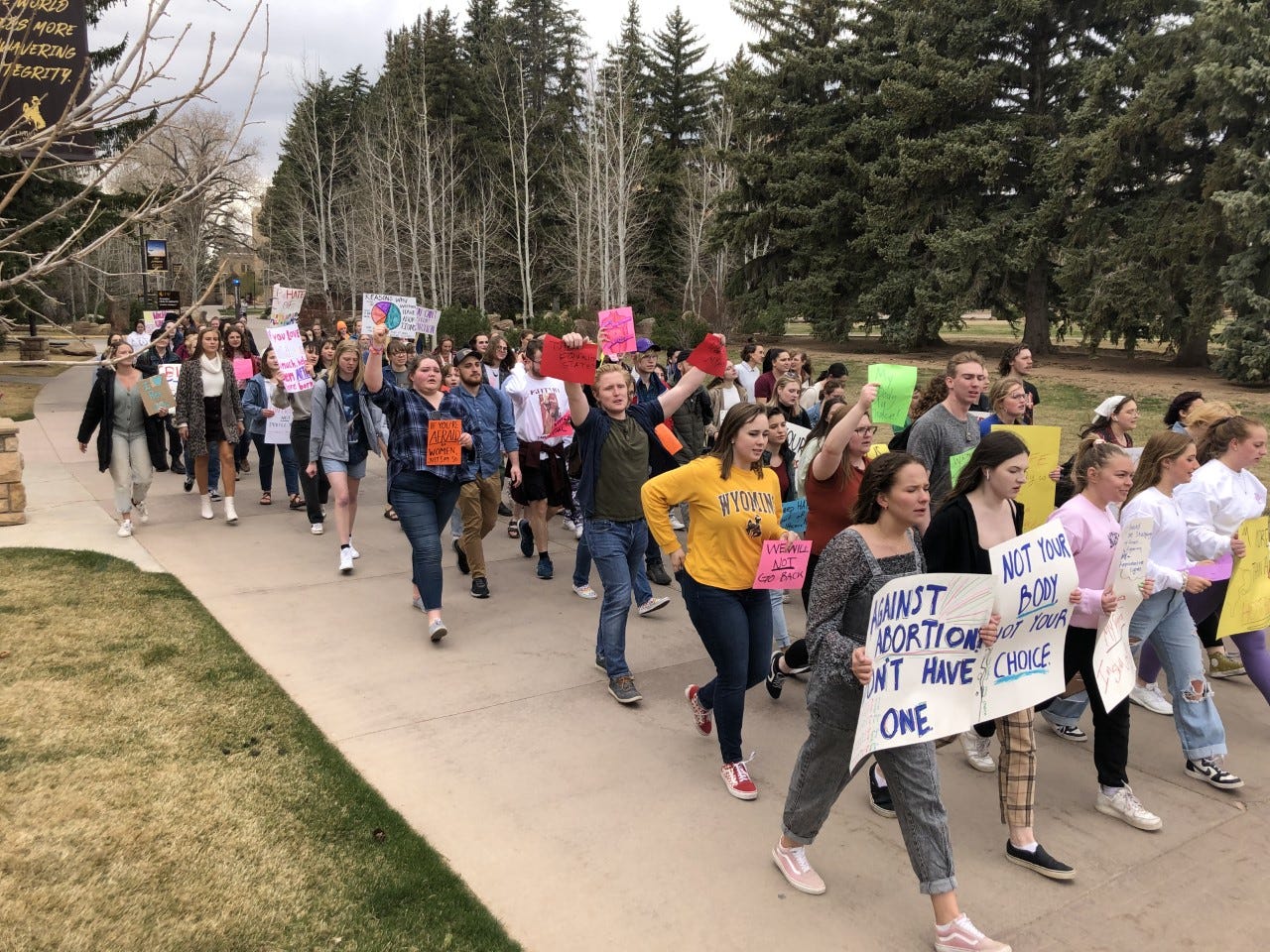 Dozens of young people carrying signs with pro-choice slogans march toward the camera on a wide sidewalk in front of tall evergreen trees.