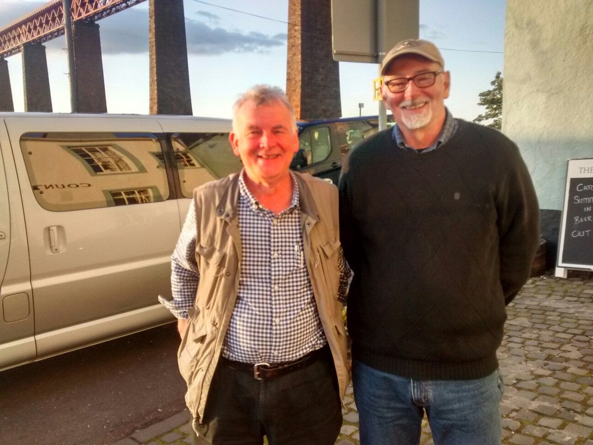 Photograph of Ken MacLeod and John Sundman, standing outside on a cobblestone patio. Immediately behind them are a long silver limousine and a black SUV. Behind the vehicles there is a high steel bridge held up by dramatic tall stone pillars. 