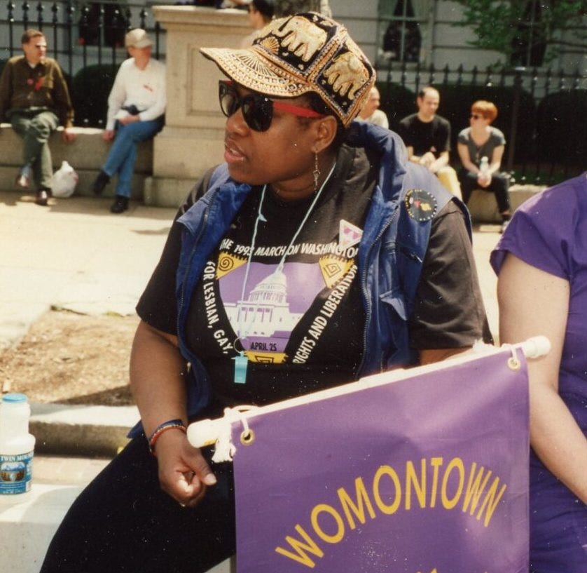 A Black woman, wearing an embroidered baseball-style hat, sunglasses, and a black t-shirt from a political march, holding a lavender banner that reads 'WOMONTOWN' in yellow letters. 
