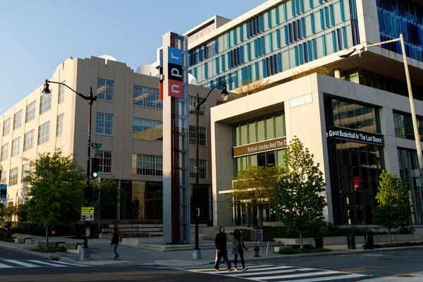 People walking in front of a large office building near a column bearing the red, black and blue NPR logo.