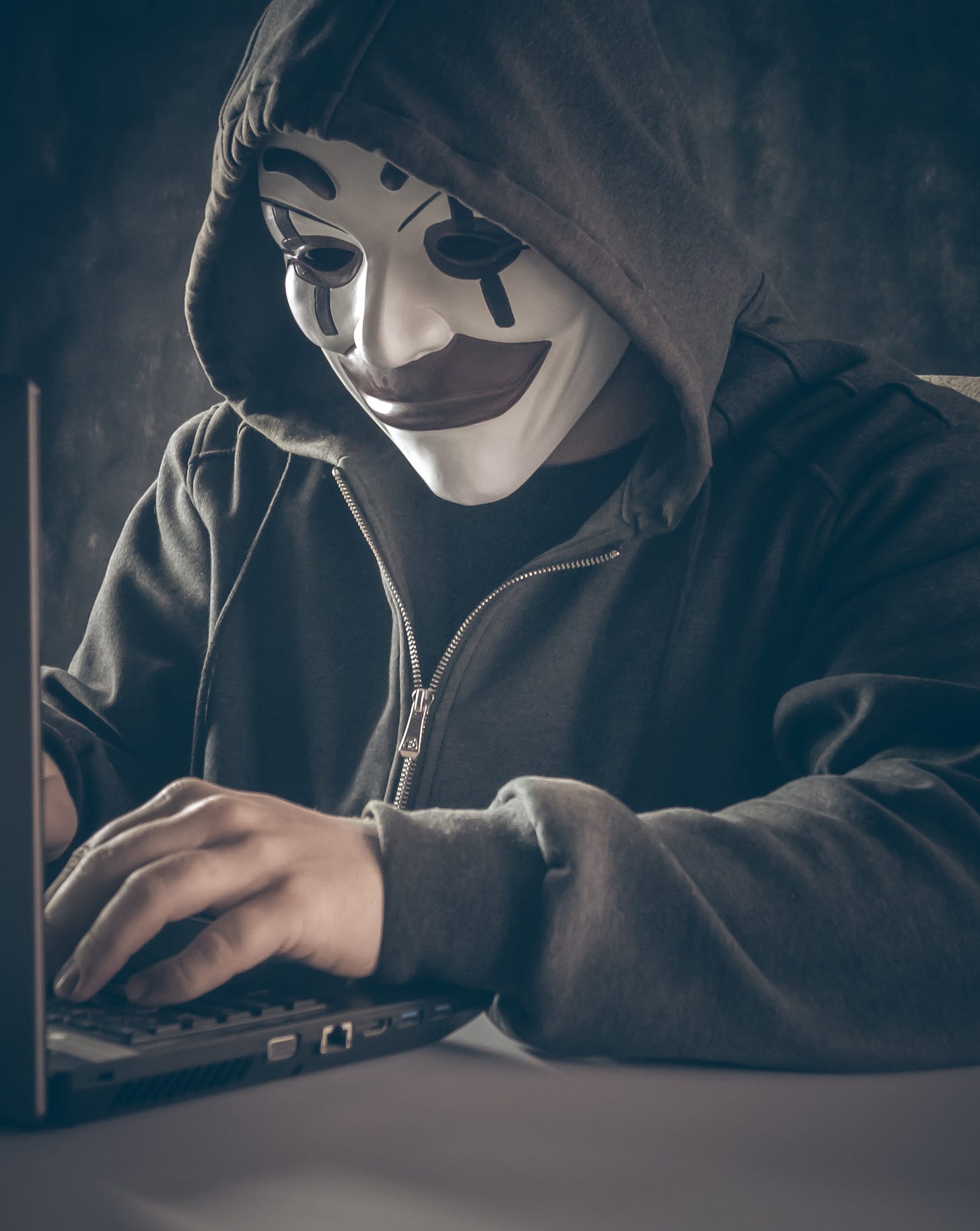 Man in pseudo-Anonymous mask with black hoodie, typing on a computer.
