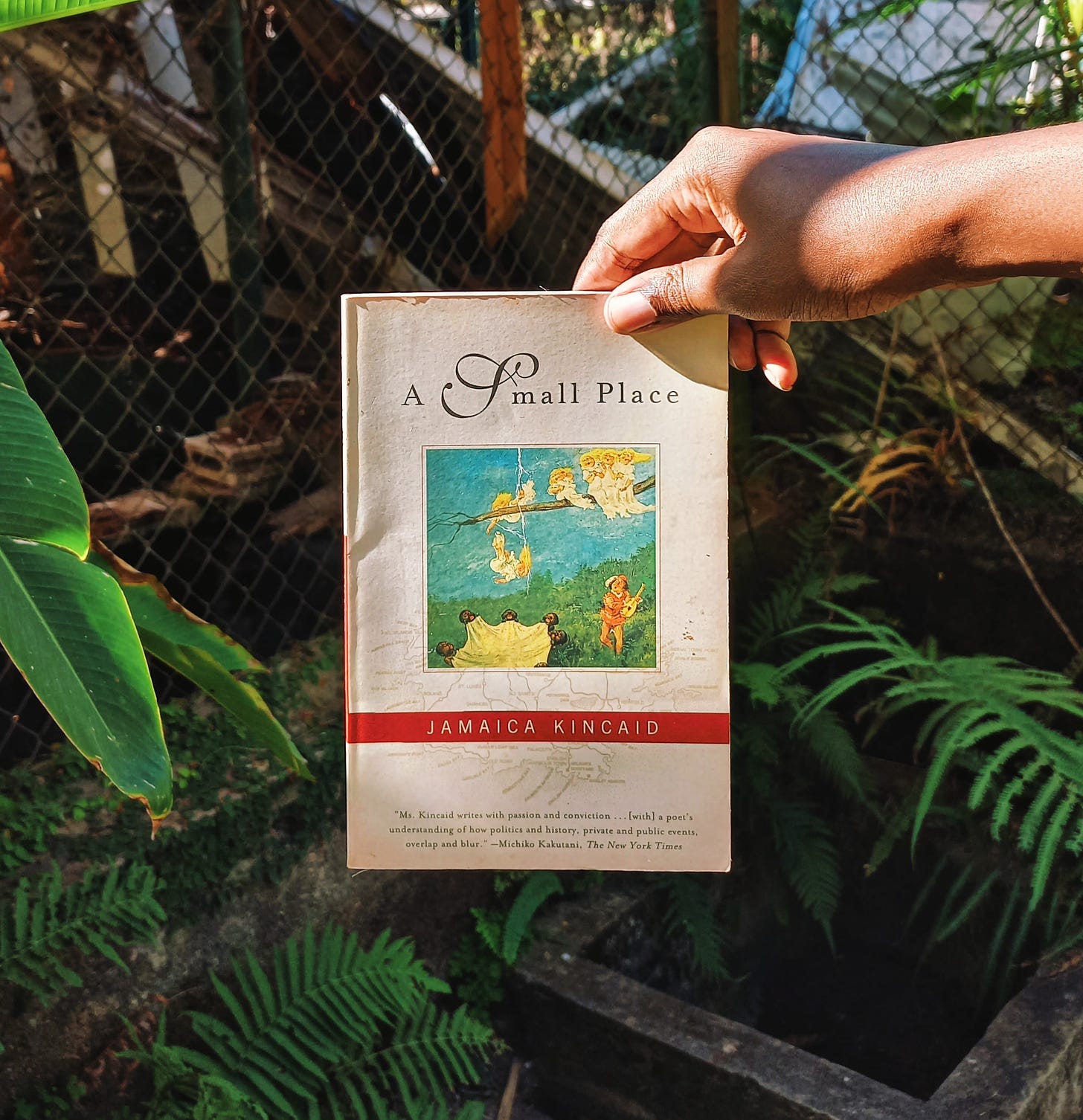 My black hand holds a paperback copy of A Small Place with plants and part of a wired enclosure in the background.