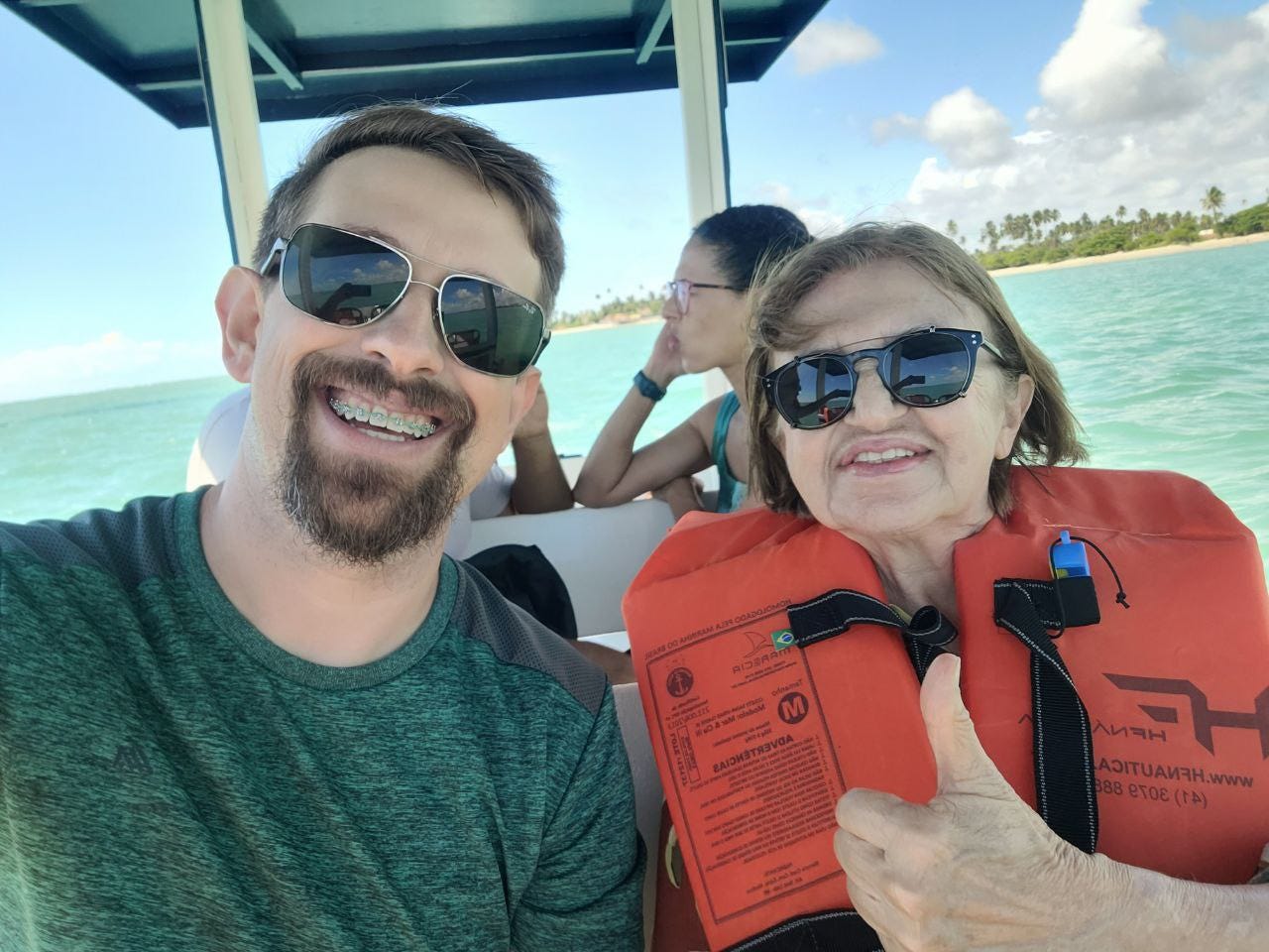 Em cima de um pequeno barco aberto, do qual apenas vemos um pedaço do teto e de duas vigas, eu e minha mãe sorrimos. Ela tem cabelo castanho claro e usa um colete de proteção laranja, enquanto faz um joia com a mão. Eu estou com camiseta verde, uso cavanhaque e sou loiro. Ambos somos brancos e usamos óculos escuros.