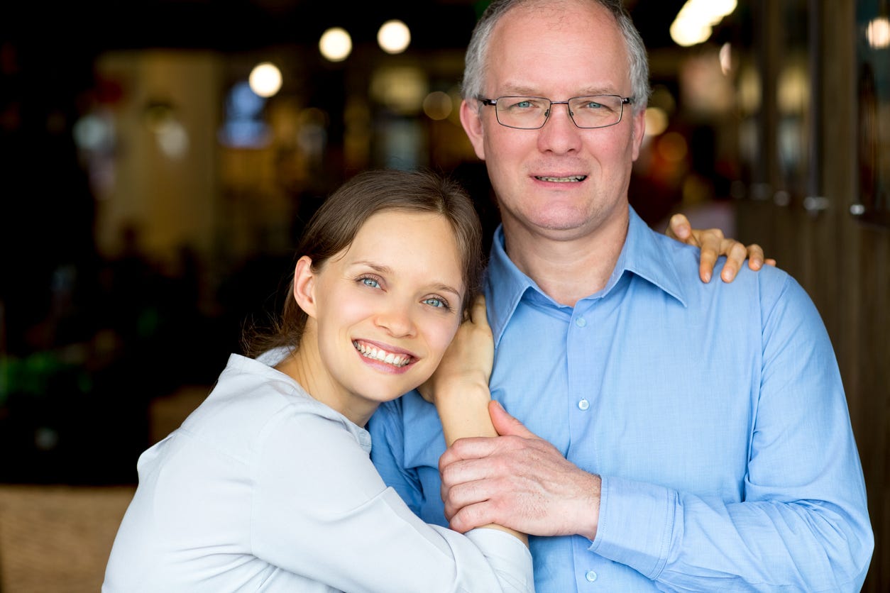 Smiling, young white woman resting her face on right shoulder of smiling, aging, white, balding man with glasses.