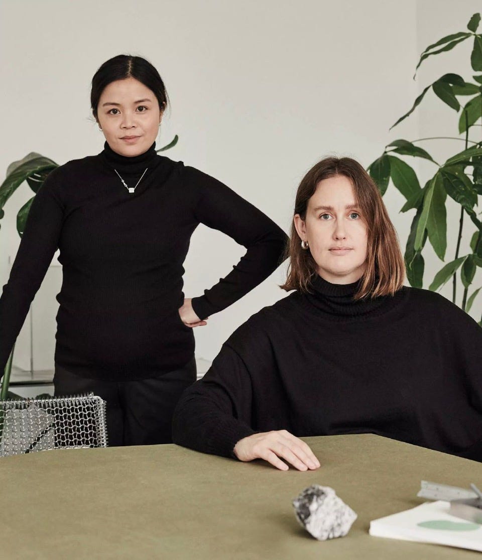 Photograph of one woman sitting down at a desk with another woman standing behind her, and plants in the background