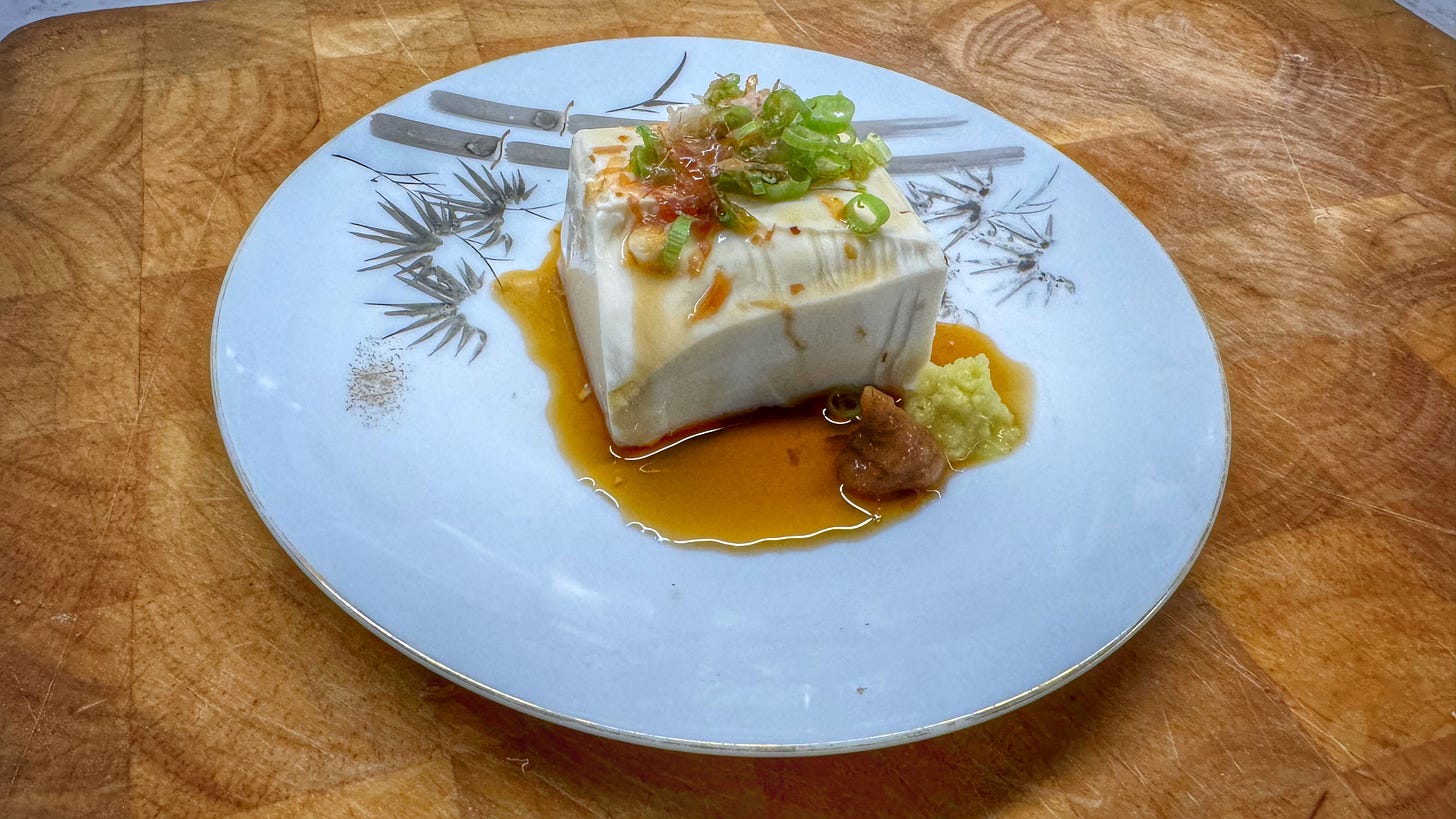 A plate of hiyayakko, with a block of tofu topped with sliced green onion and katsuo flakes. Ponzu has been poured over it and there are piles of ginger and plum paste on the plate. 