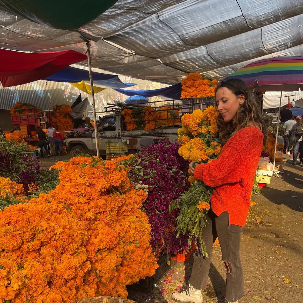 A woman in a red jumper holds yellow flowers infront of a flower market stall full of cut flowers in orange and red.