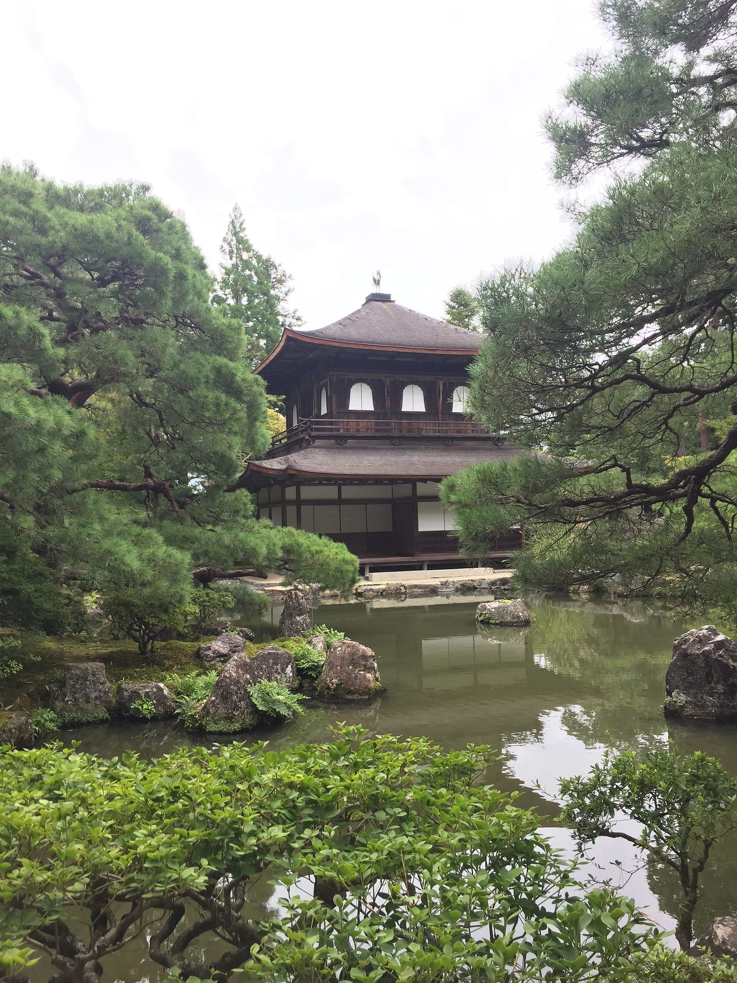 A two-storey wooden temple, surrounded by trees, is viewed from across a large pond with feature stones.