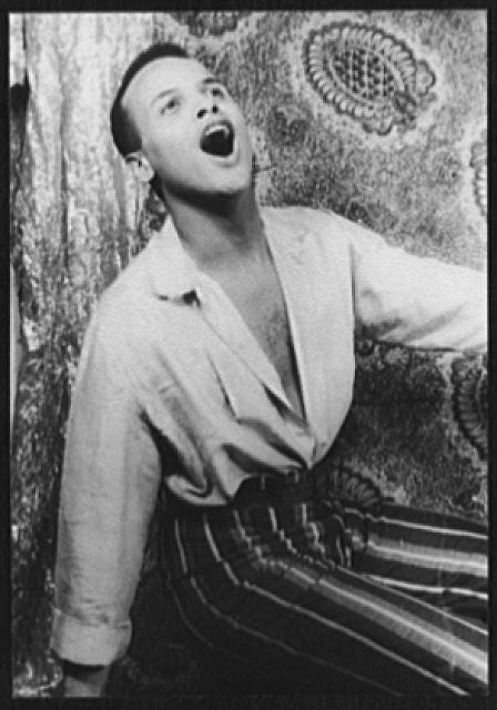 A photograph of Harry Belafonte. His head is leaning back and his mouth is open singing. He has his shirt unbuttoned all the way down and tucked in. He is sitting wearing striped pants.