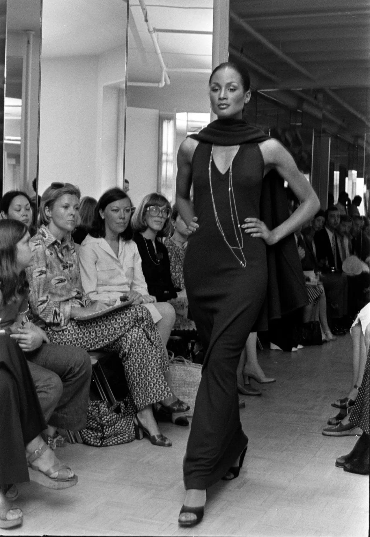 A model clad in clinging V-neck cashmere dress and scard walks down a runway as members of the fashion press and house clients look on. 