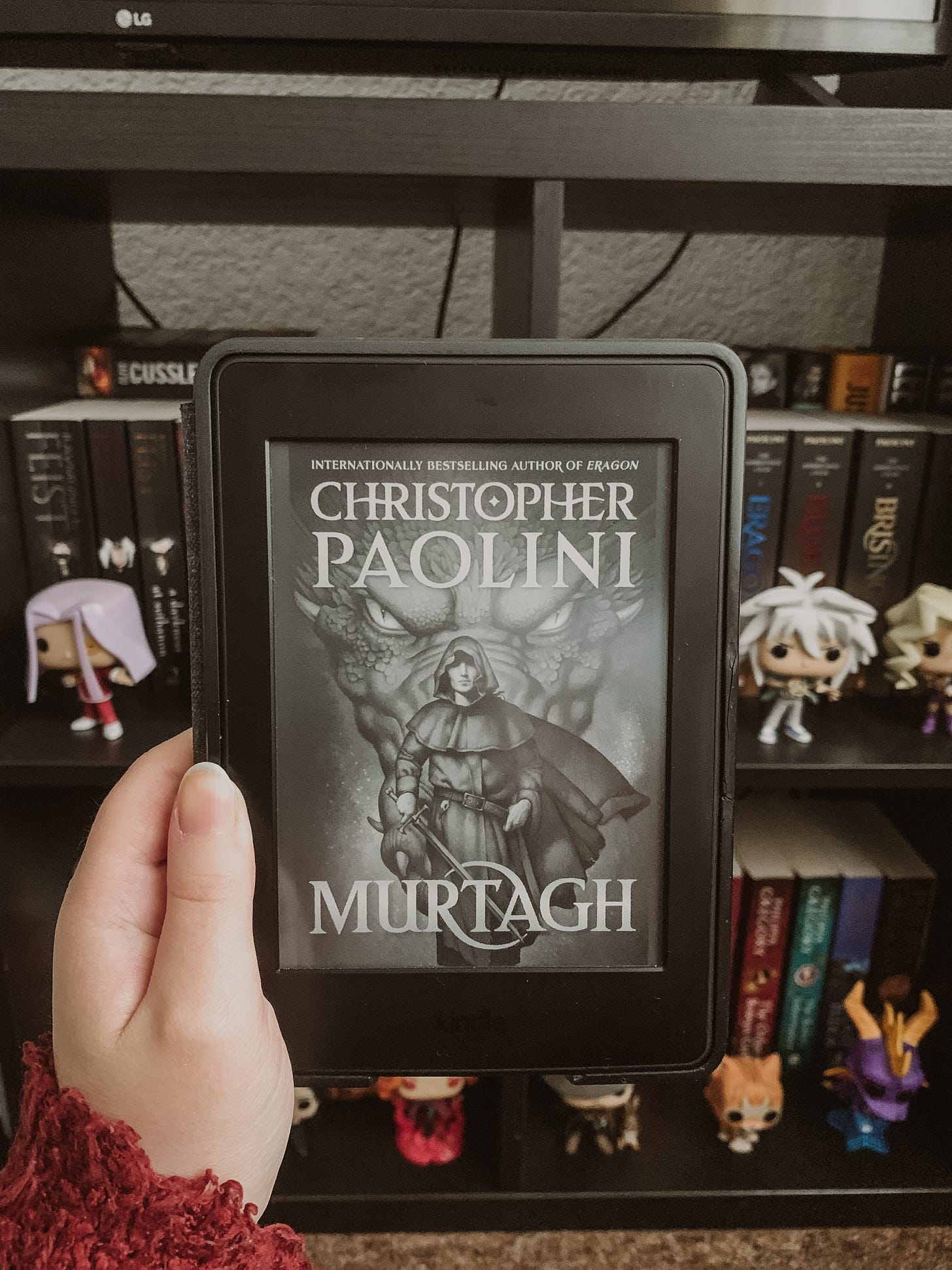 A black kindle being held up in front of a bookcase, with the fron cover of Murtagh by Christopher Paolini on the screen