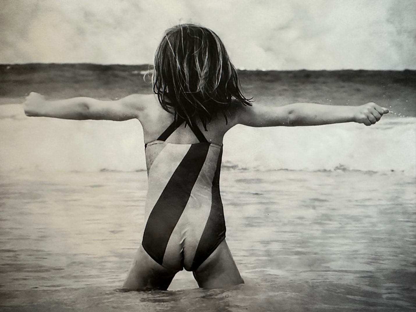 Small girl stands defiantly with arms outstretched facing incoming ocean waves