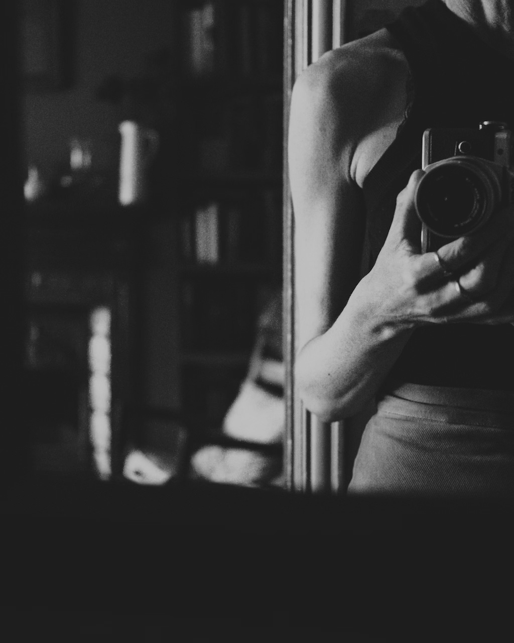 A woman is partly seen taking a photograph into a mirror, with a room visible in the background (in black and white)