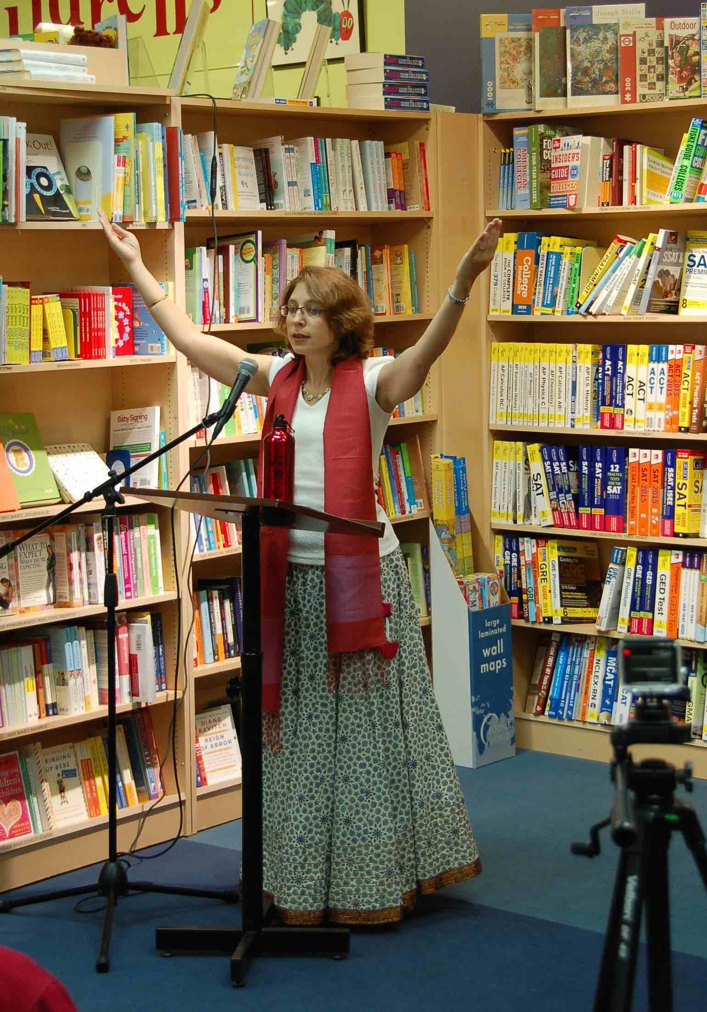 Woman in skirt and white shirt with a red scarf, at a podium, with arms raised, surrounded by bookcases.