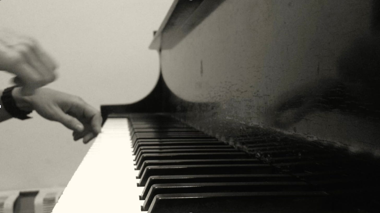 A black-and-white image shows a closeup of a piano keybed at right and Tara's hands at left. The left hand is at rest, barely touching a key, and the right hand is in motion above the keys.
