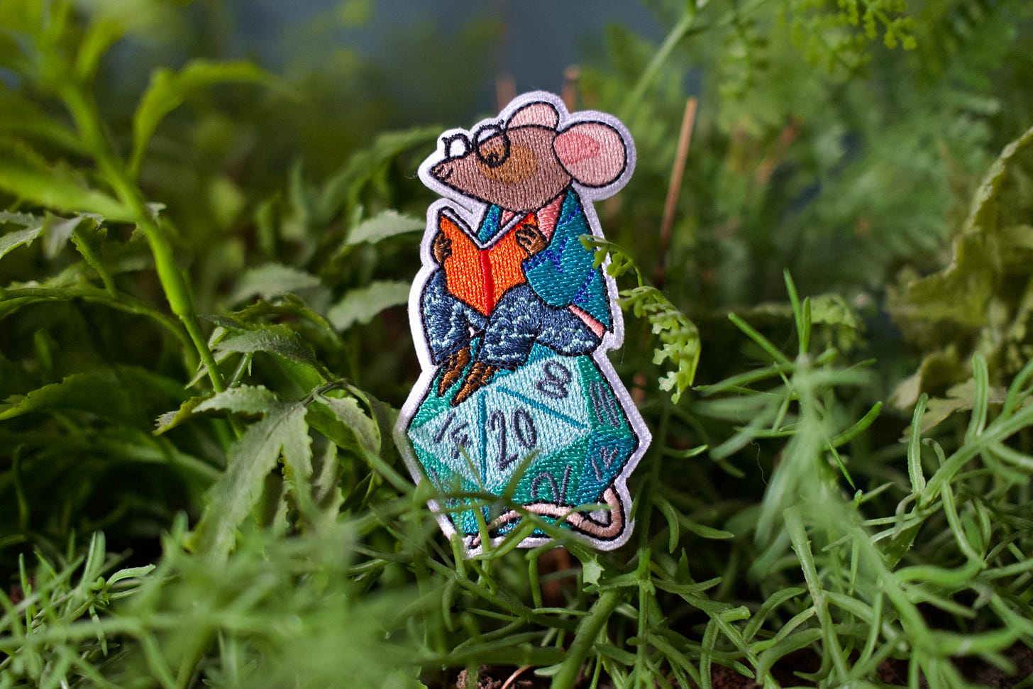 A photo of a fabric patch. The patch is a mouse sitting on a twenty-sided dice.