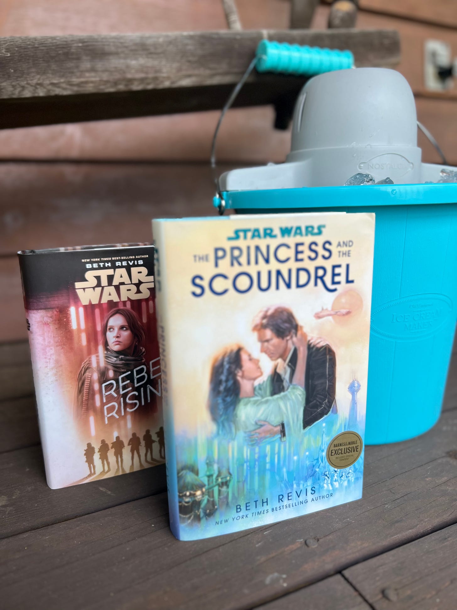 Copies of Rebel Rising and The Princess and the Scoundrel on a wooden porch in front of a blue ice cream maker
