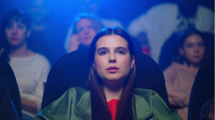 Young woman with black hair, red top, and green jacket in crowded theater watching a film with concern on her face.