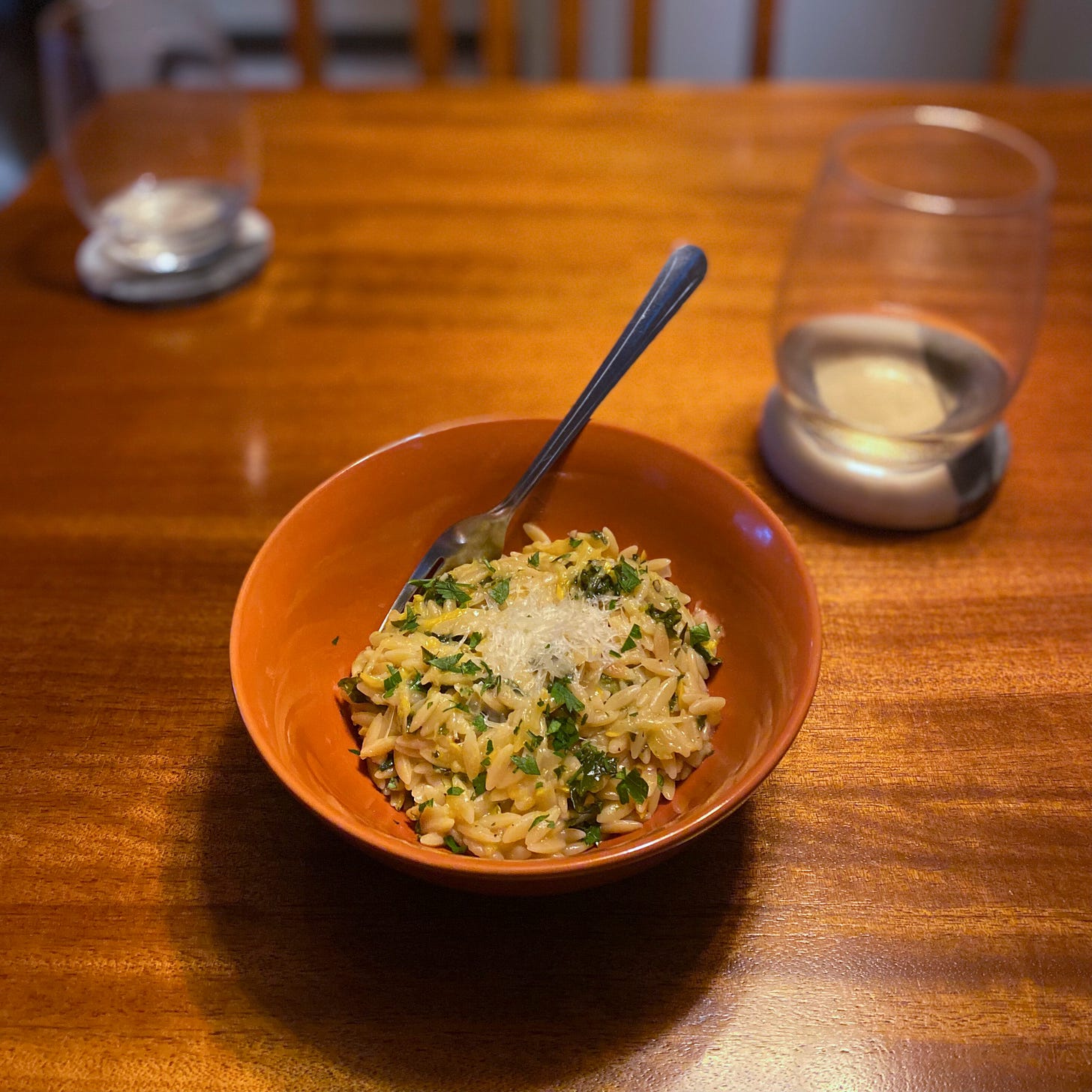 An orange bowl of the orzo described above, with pieces of kale and parsley visible throughout. Parmesan is grated into a little pile on top, and a fork rests at the back of the bowl. On a coaster to the right is a glass of white wine.