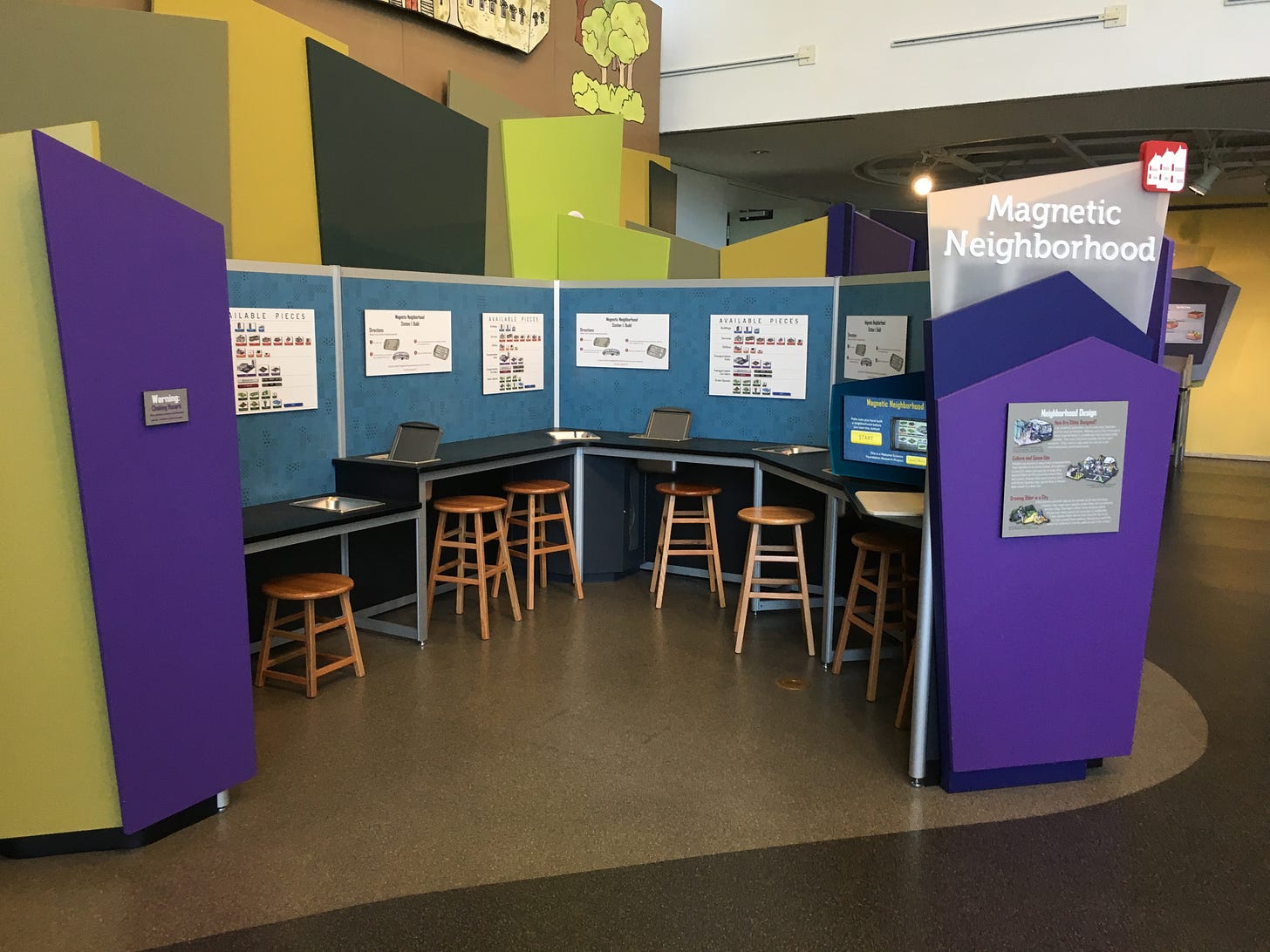 A U-shaped table enclosed by blue and purple walls, with six stools at the table. On the table are bins holding metal baking sheets. On the walls are posters that describe what magnets people can use on the trays.