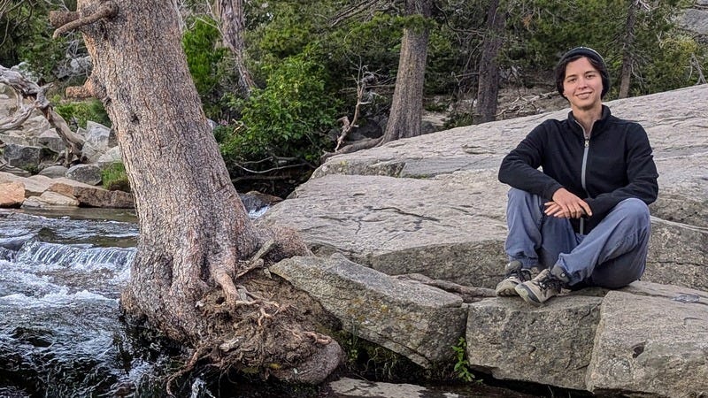 Rey, a white non-binary person, sitting on a rock next to a flowing stream