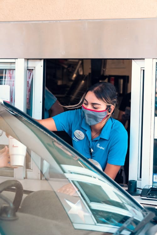 Free A fast-food drive-thru employee wearing a mask serves a customer. COVID-19 safety measures in place. Stock Photo