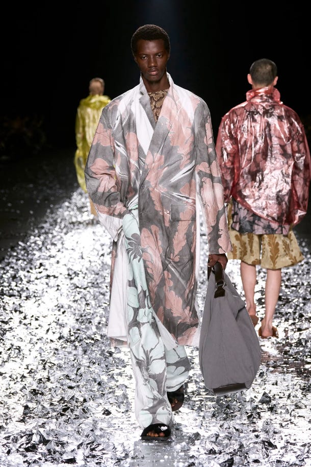 Black male on a catwalk wearing a faded pink and grey floral trenchcoat and similarly patterned bright wide legged trousers.