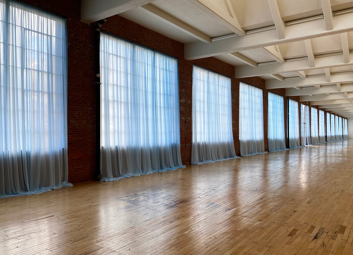Installation view of Felix Gonzalez-Torres “Untitled” (Loverboy), 1989, at Dia Beacon. A series of sheer light-blue curtains are installed along the long outer wall of a huge industrial-scale room with a wooden floor.