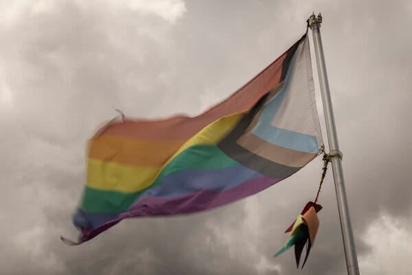 A rainbow-colored pride flag waving on a flagpole against a cloudy sky.