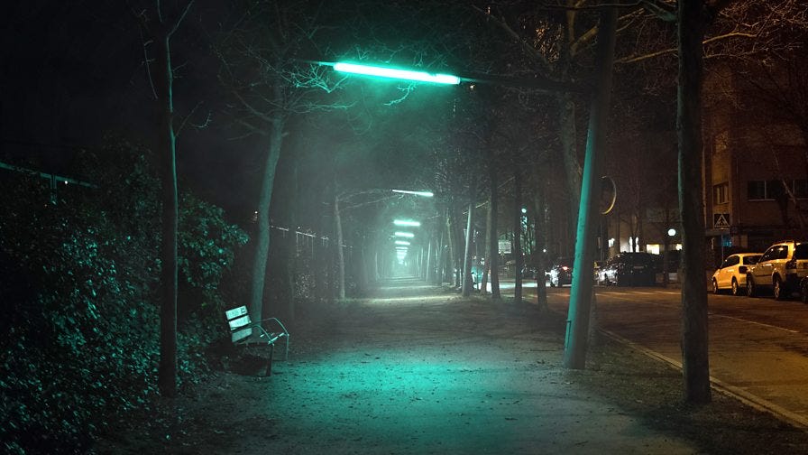 artwork van Lights out. Een foto van een straat, 's avonds. In het midden groen, onheilspellend licht dat van lantaarns komt en een pad vormt