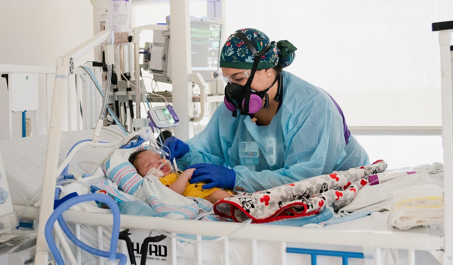 Photo of a nurse wearing an elastomeric respirator treating a pediatric COVID-19 patient in the ICU hooked up to a ventilator