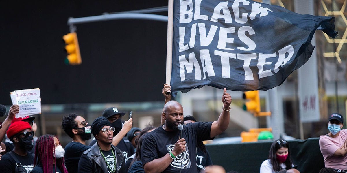 A man delivers a speech to a crowd in front of a large Black Lives Matter flag at a protest in New York