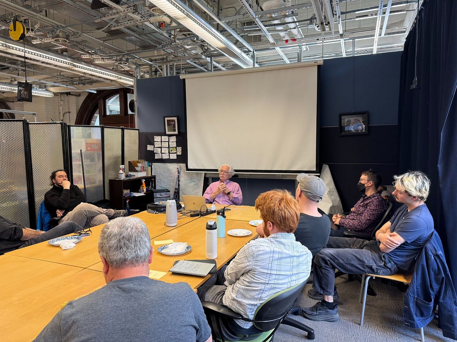Terry Winograd speaks to a small group seated around a conference table in a casual meeting room with a projector screen behind him. Terry Winograd speaks to a small group seated around a conference table in a casual meeting room with a projector screen behind him.