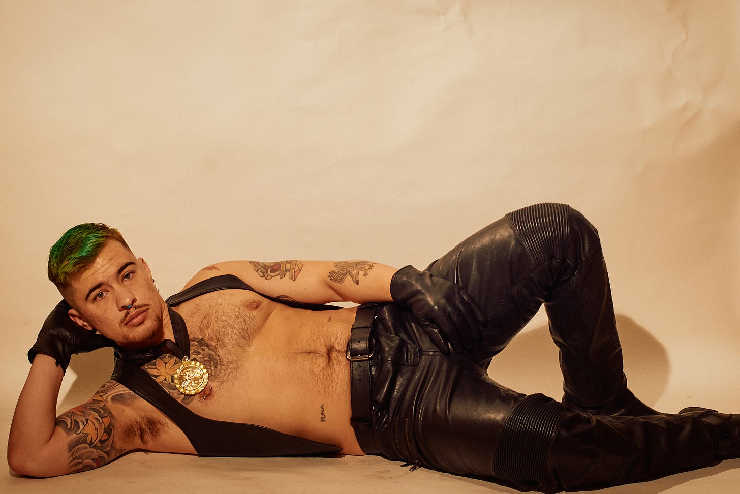 A person reclines on their side, dressed in black leather pants, gloves, and a harness adorned with a gold medal. Their pose is relaxed yet striking, highlighting their tattoos and green hair with a soft, neutral backdrop.