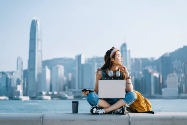 Beautiful young Asian woman sitting cross-legged by the promenade, against urban city skyline. She is wearing headphones around neck, using smartphone and working on laptop, with a coffee cup by her side. Looking away in thought. Lifestyle and technology Beautiful young Asian woman sitting cross-legged by the promenade, against urban city skyline. She is wearing headphones around neck, using smartphone and working on laptop, with a coffee cup by her side. Looking away in thought. Lifestyle and technology hong kong woman stock pictures, royalty-free photos & images