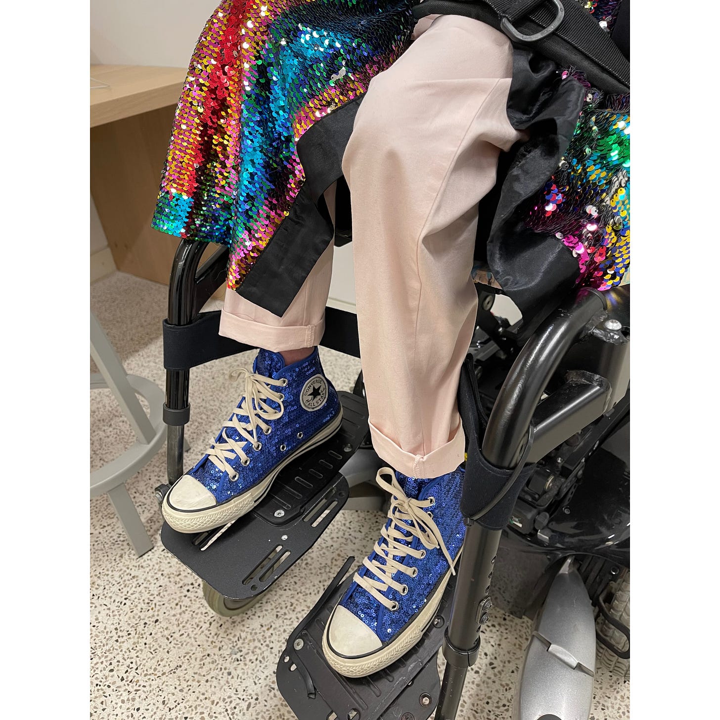 IMAGE DESCRIPTION 4: Sulaiman sitting in his power wheelchair in the greenroom at The Design Museum after his TED talk in front of a poster on the wall. He is wearing his white french cuff, witty cufflinks, his rainbow coloured sequinned kimono, Custom Cripjoy badge, Silver Nkonsonkonson Adinkra pendant via Ahima Jewelry representing the importance of family (whether by blood or bond) to me. Sulaiman is also wearing his Silver signet ring via Vitae London engraved with “Janed” on the top representing Camp Janed and the wisdom of Disabled people, his Authentic Ghanaian beads bracelet PokuDots and Michael representing his accompliceship for the #BlackLivesMatter movement, his stylish pink chinos, his blue sequinned converse shoes, and a “#BlackDisabledLivesMatter” face mask via Jennifer White-Johnson.