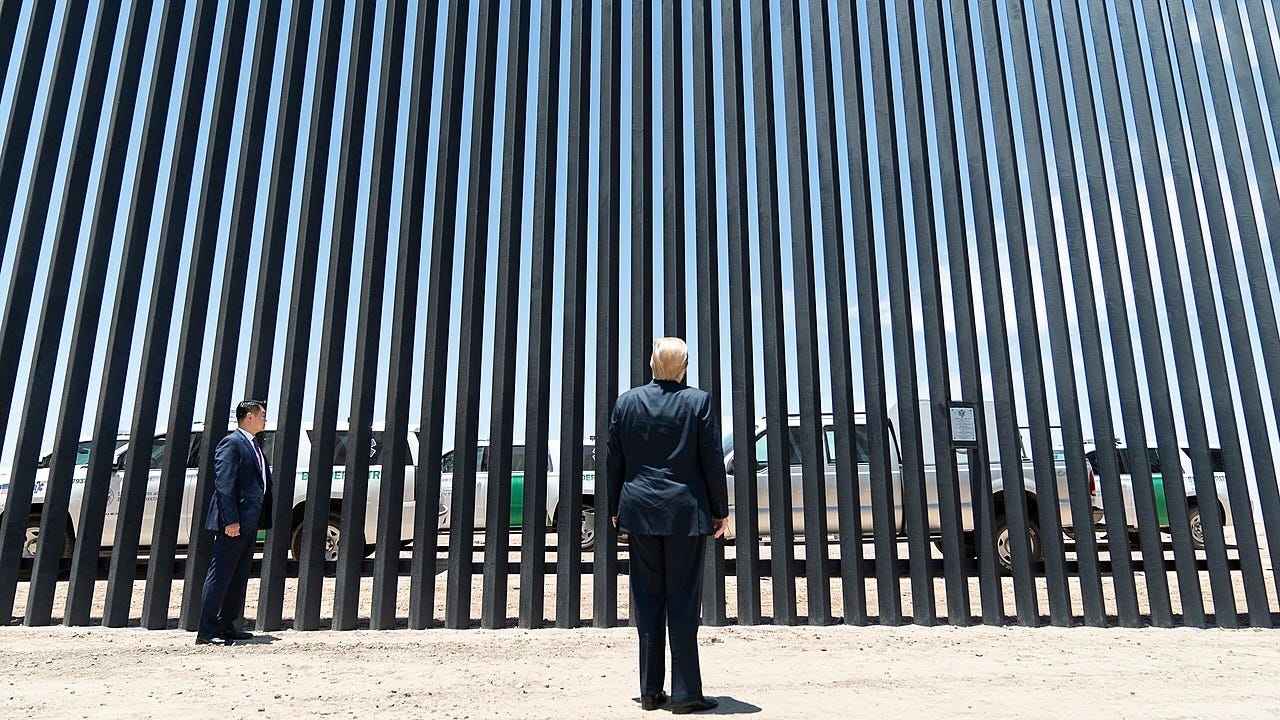 Donald Trump stands in a blue suit with this back to the camera looking at the slats of a section of border wall.