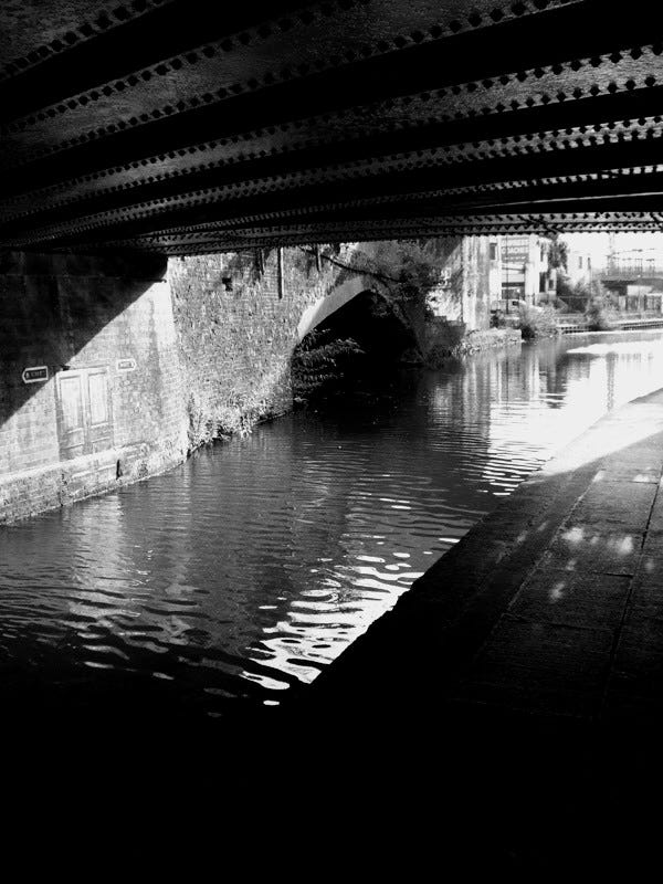 A black and white image of an iron bridge above the Regent's Canal in East London, with a narrow towpath.