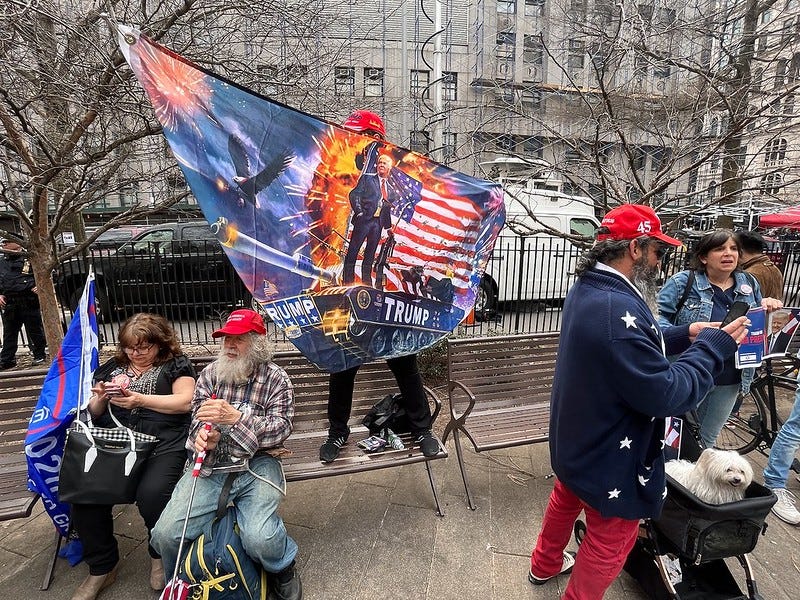 A photograph of a group of protestors outside the Manhattan courthouse on the day of Trump's arraignment. In the center, a man is waving a banner that features a portrait of Trump standing on top of a tank (labeled "TRUMP"), holding a weapon and with an American flag behind him. There are bald eagles and explosions.